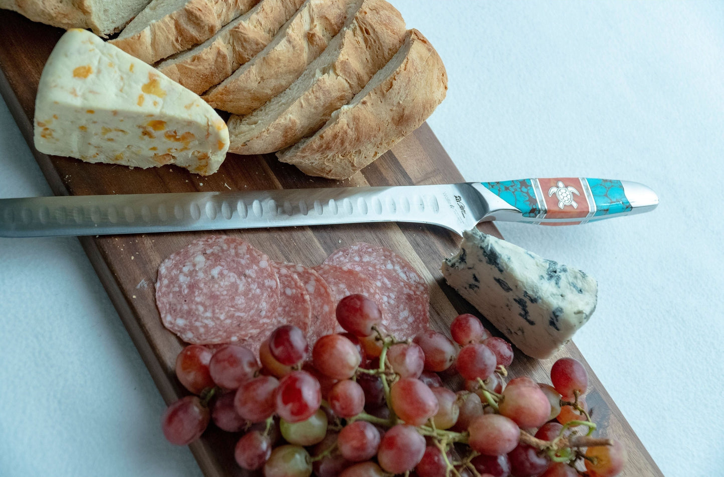 Wooden cutting board with a knife, bread, cheese, salami, and grapes on a white background