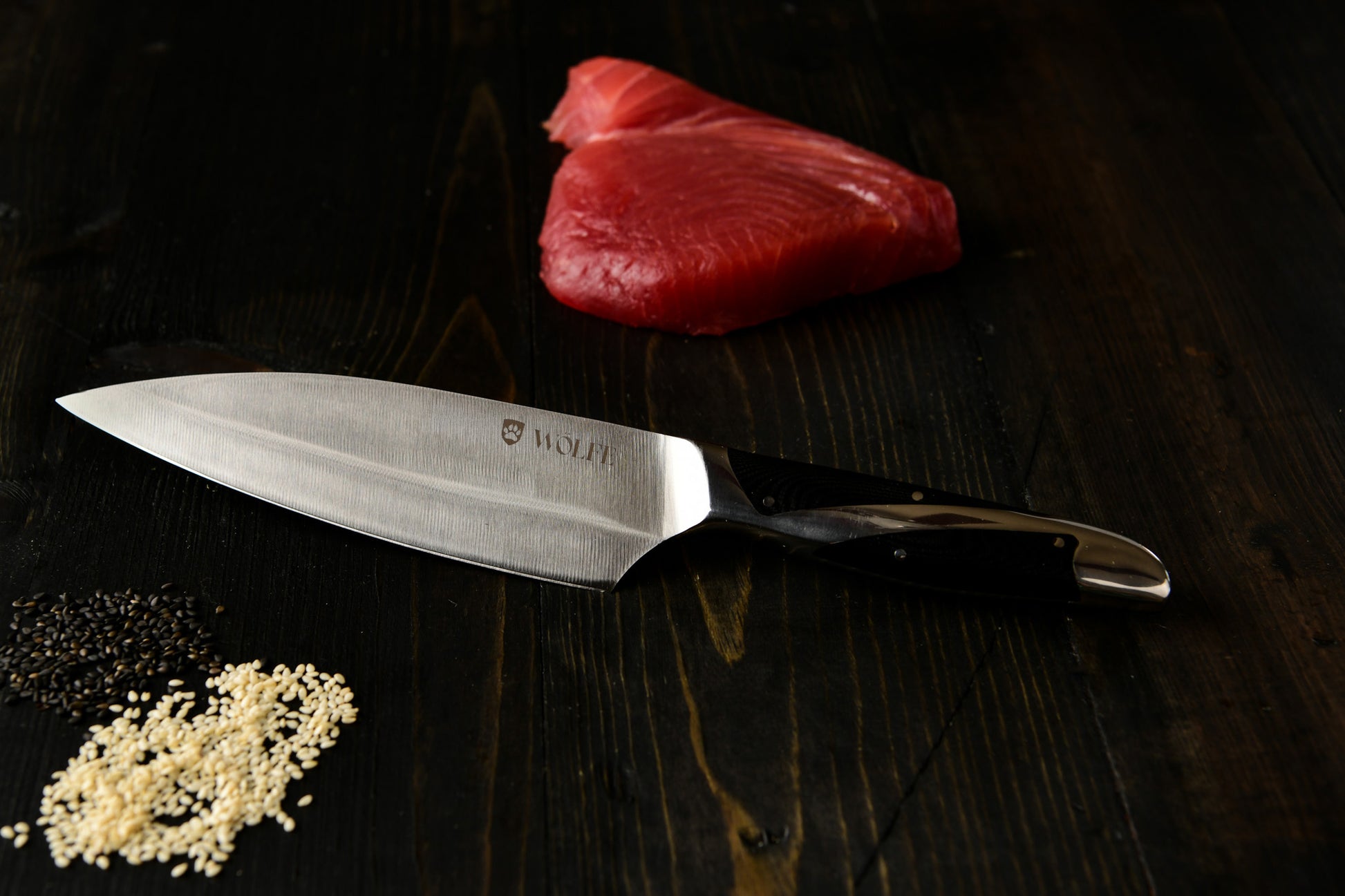 Chef's knife with the Wolfe logo on a dark wooden surface next to raw fish and sesame seeds.