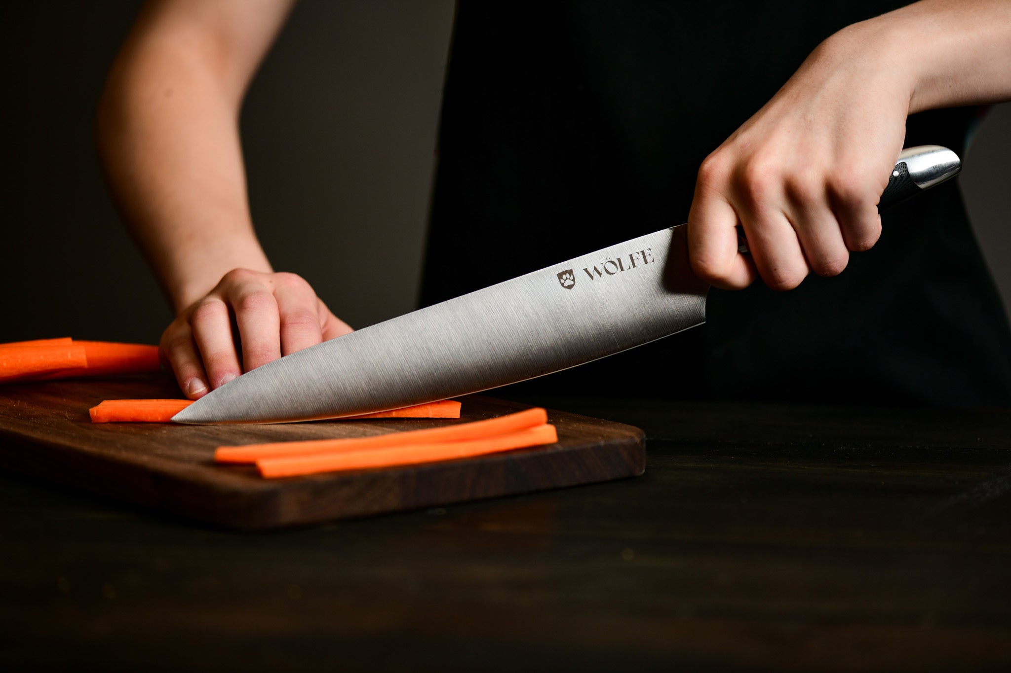 Person using a Wolfe knife to cut carrots on a wooden board with a dark background