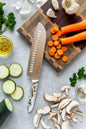 Knife on a wooden cutting board with sliced carrots, zucchini, and mushrooms on a gray surface.
