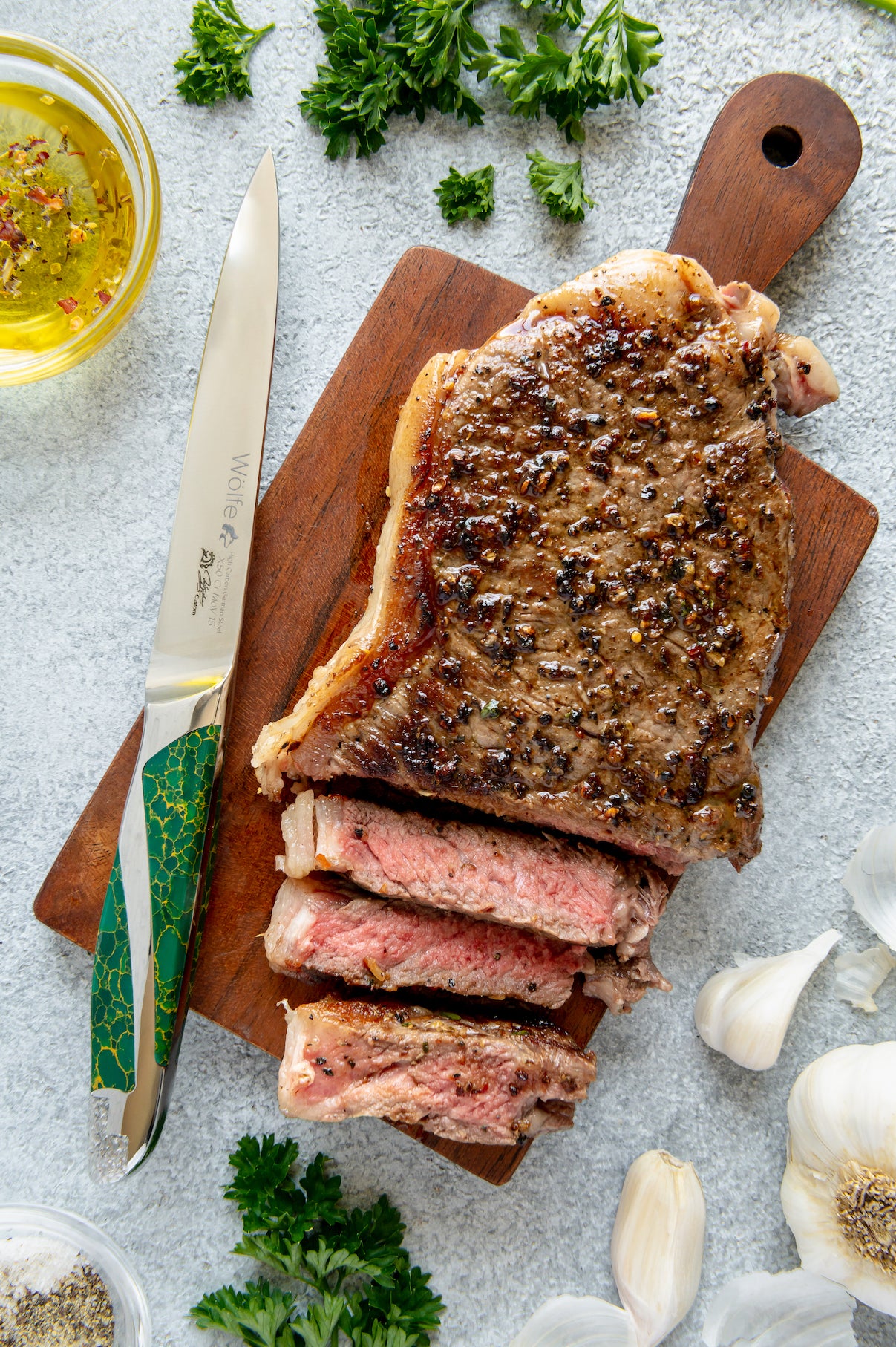 Sliced steak on a wooden cutting board with a steak knife, herbs, and garlic on a light gray surface