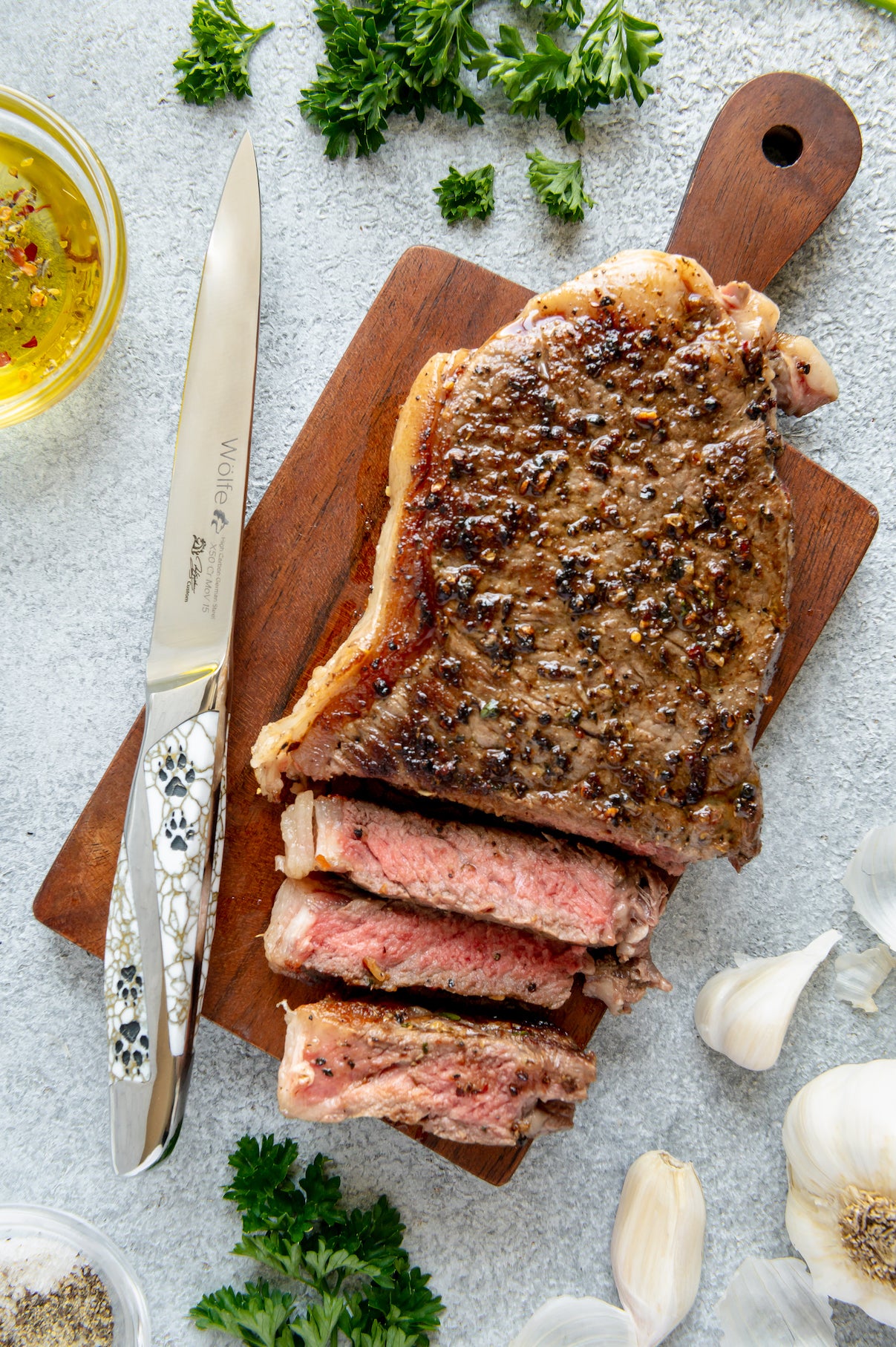 Sliced steak on a wooden cutting board with a knife, herbs, and garlic on a light gray surface