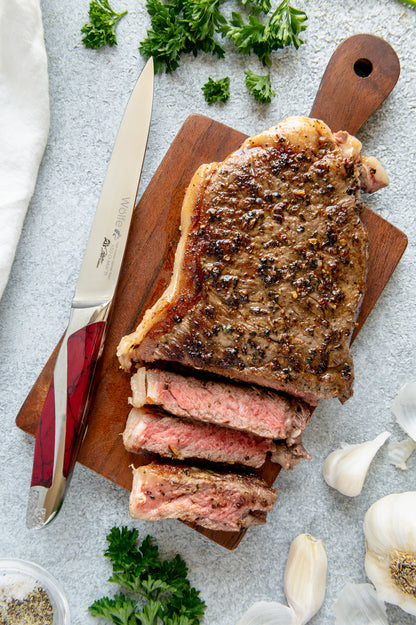 Sliced steak on a wooden cutting board with a knife, herbs, and garlic on a light gray surface