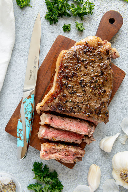 Sliced steak on a wooden cutting board with a knife, herbs, and garlic on a light gray surface