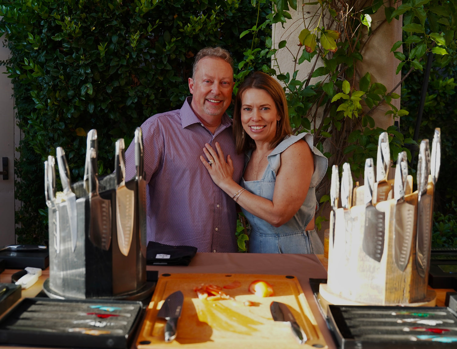 Man and woman posing together with a display of knives and cutting boards.