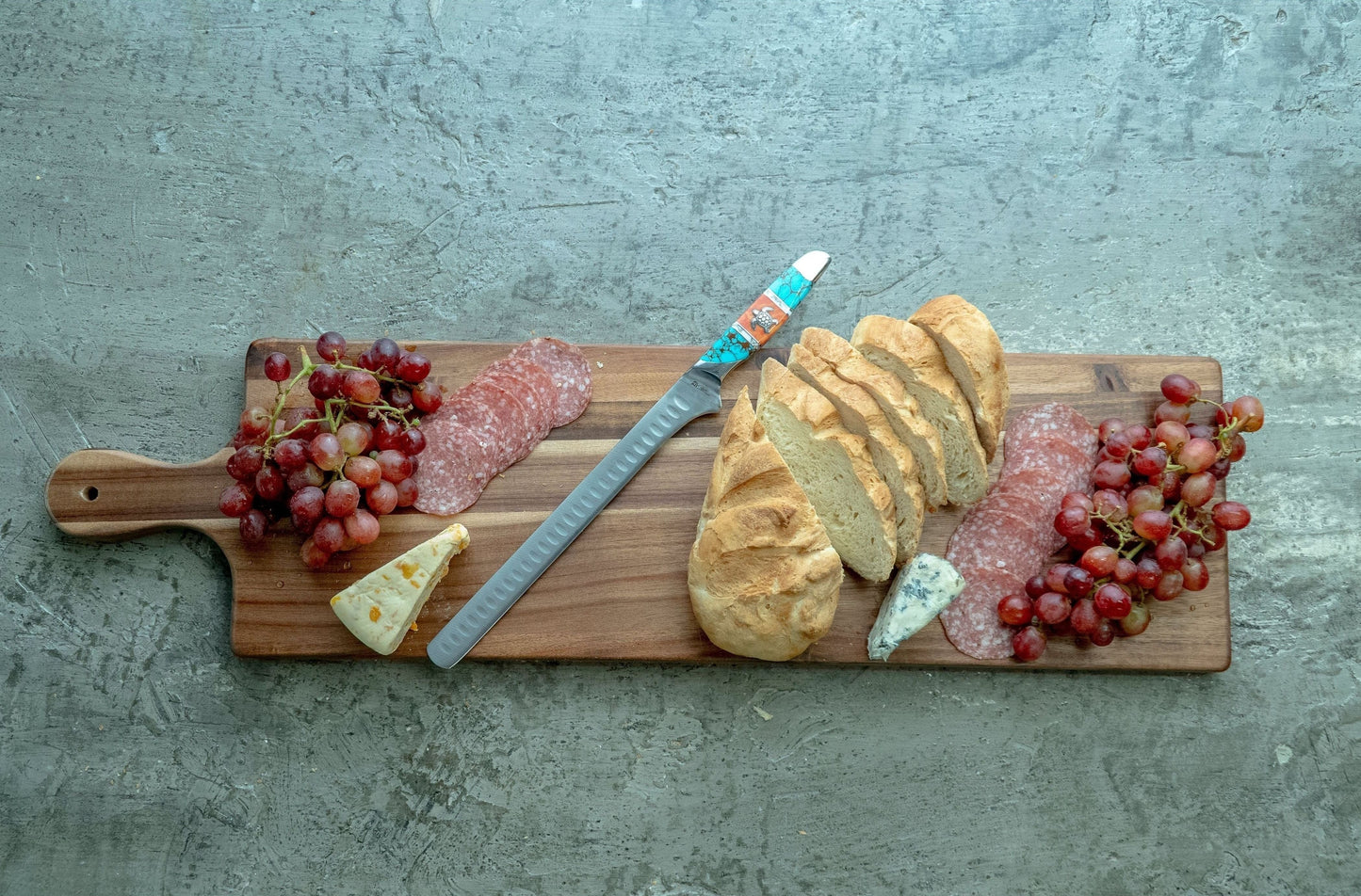 Wooden cutting board with a knife, bread, salami, cheese, and grapes on a concrete surface