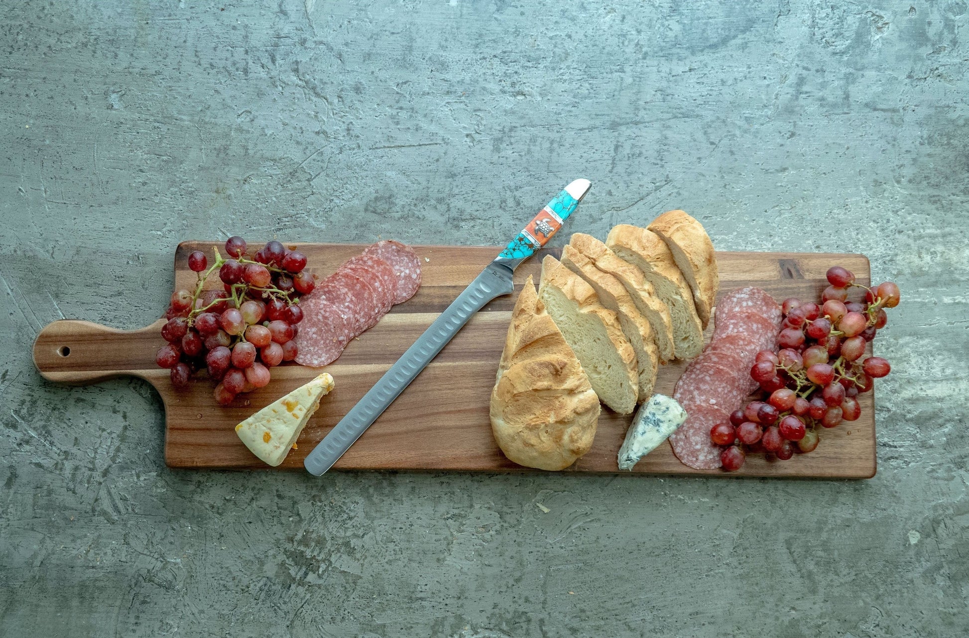Wooden cutting board with a knife, bread, salami, cheese, and grapes on a concrete surface