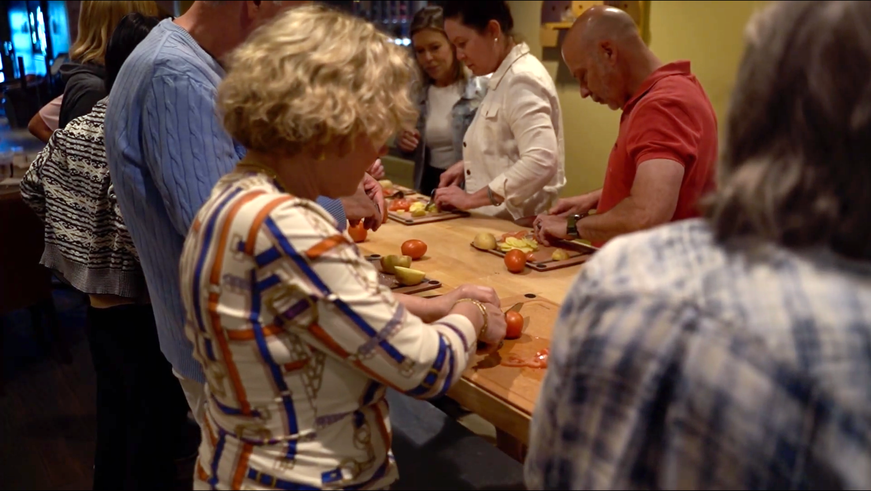 A group of people gathered around a wooden table in a kitchen or dining area, preparing food and cutting ingredients. The scene is warmly lit with a cozy indoor atmosphere.