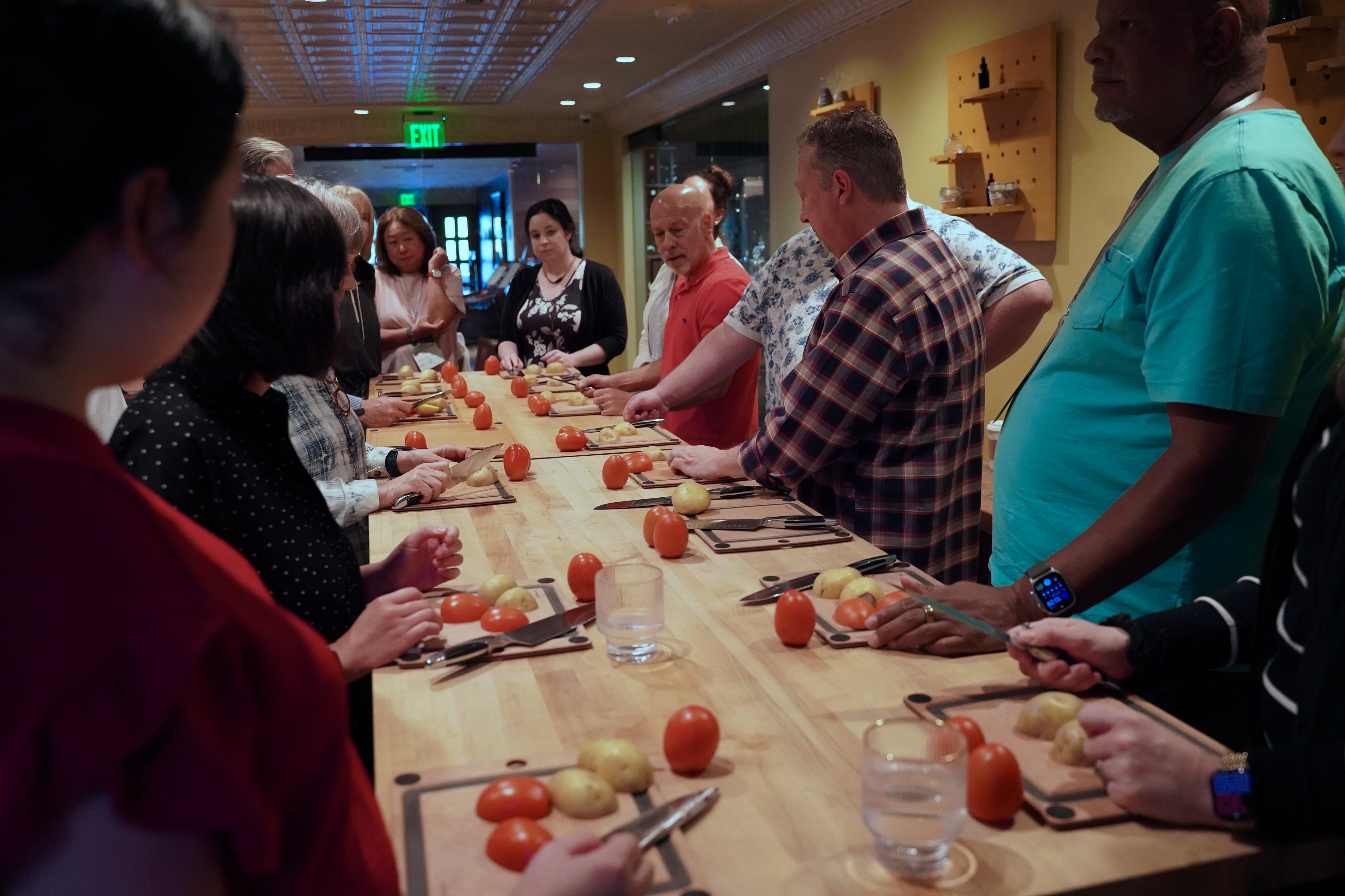 A group of people standing and sitting around a long wooden table in a kitchen or training space, preparing tomatoes and potatoes on cutting boards with knives. Several faces are blurred for privacy.