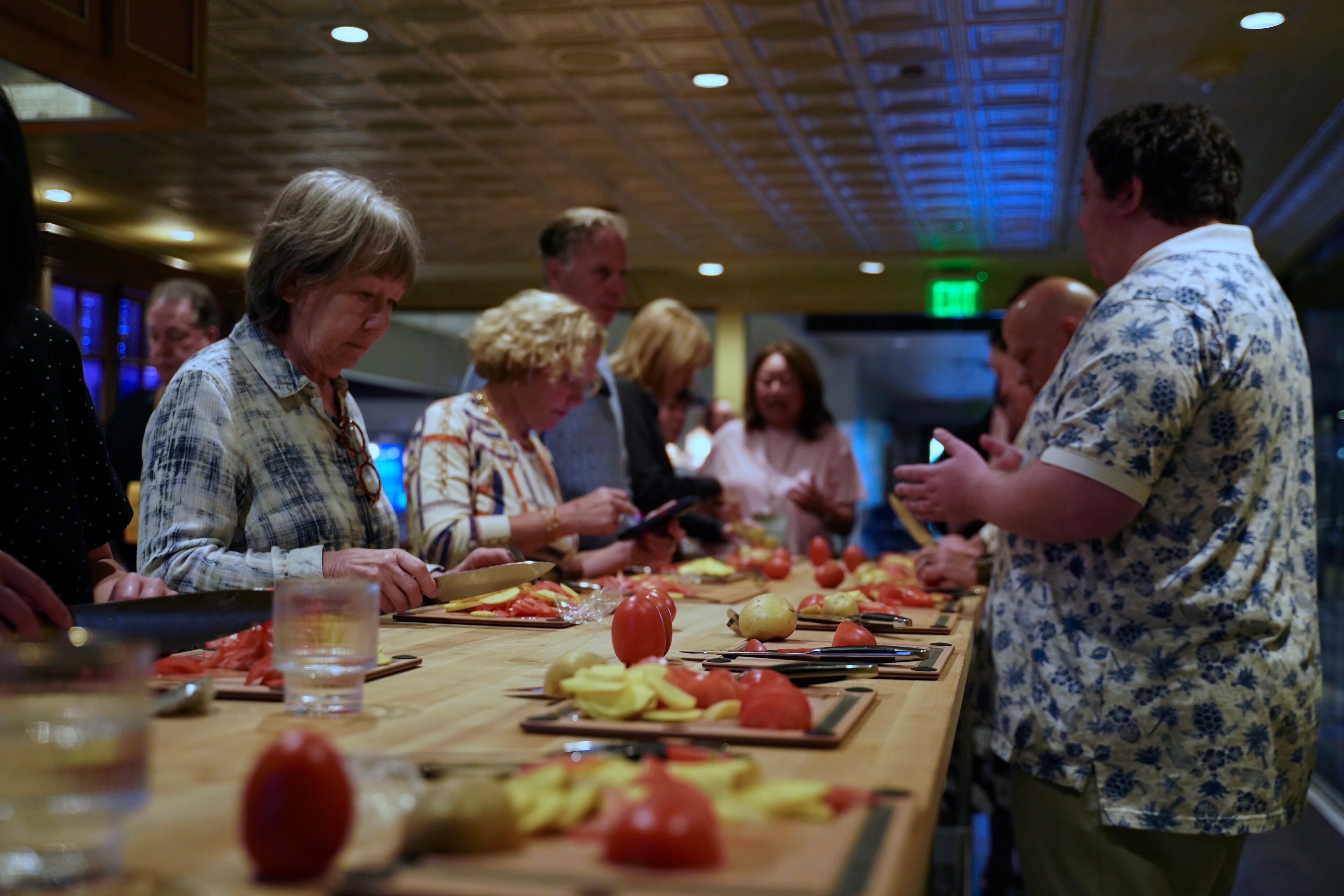 A group of people gathered around a long wooden table in a kitchen or dining space, preparing food with tomatoes, potatoes, and cutting boards under warm overhead lighting.